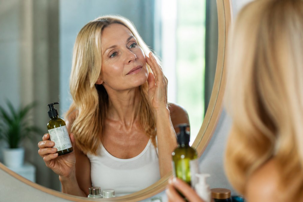 Woman in her 50s examining her reflection in a mirror with soft natural light highlighting mature skin texture, holding a bottle of organic skincare product