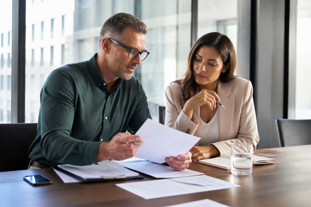 Business owner and consultant reviewing financial documents and growth metrics on laptop in bright, modern office setting with natural window lighting