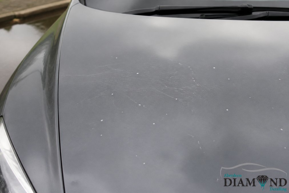Close-up of a grey car bonnet under overcast daylight showing a cluster of small white stone chips and fine spider-web swirl marks across the clear coat