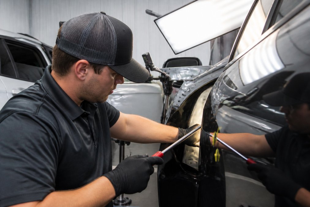 Professional technician using precision metal reshaping tools and use points on vehicle panel to remove hail dents while preserving original factory paint finish in well-lit workshop