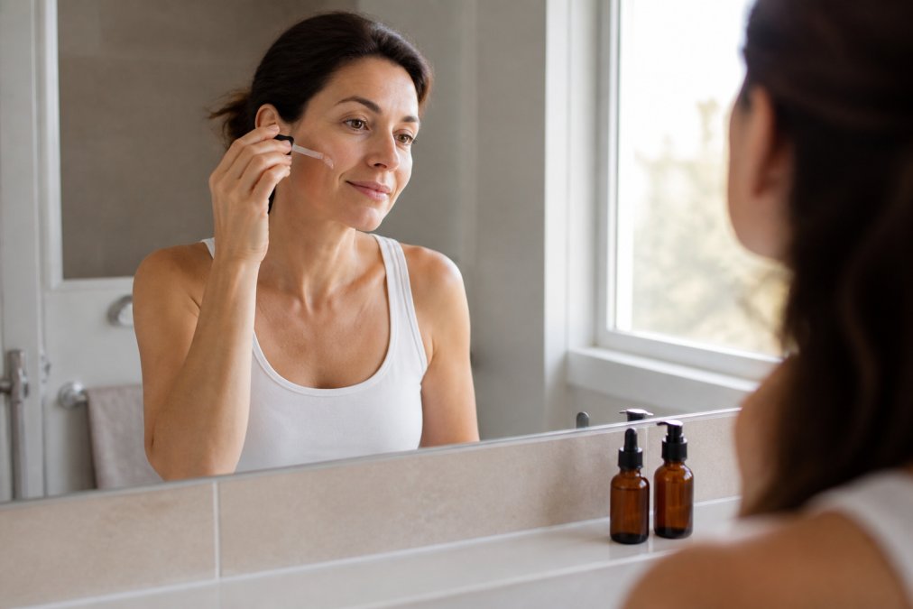 A woman in her late thirties applying a drop of serum to her cheek in a bright bathroom, with a simple two-bottle routine on the counter beside her