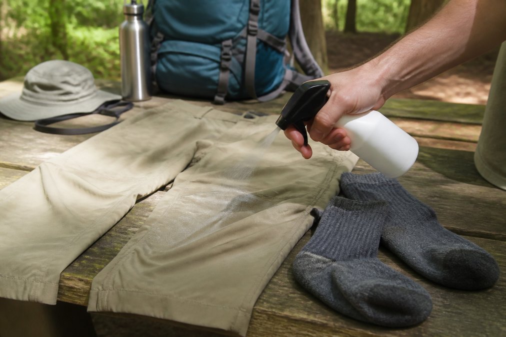 Close-up of hands spraying permethrin solution on hiking pants and socks laid out on a picnic table in natural daylight, with camping gear visible in background