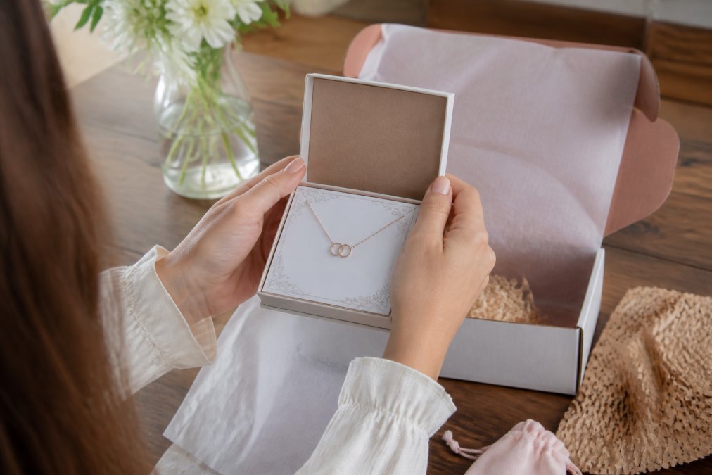 Person's hands holding a personalized necklace box with custom message card visible, examining the jewelry and packaging materials in natural lighting