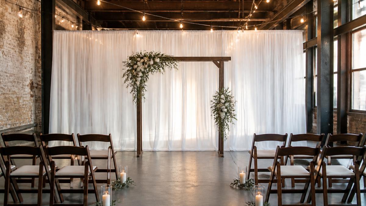 A small wedding ceremony setup in an industrial venue with white sheer voile draping forming a backdrop behind a wooden arch, two short rows of chairs, warm string lighting overhead