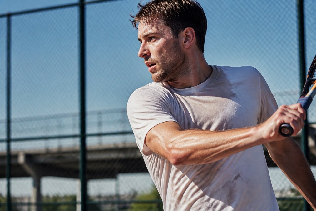 Tennis player mid-rally wearing a sweat-soaked shirt.