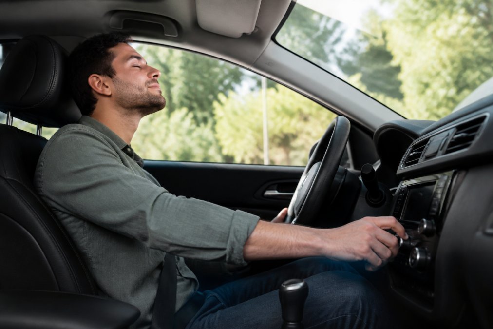 A person sitting in a car driver's seat with eyes closed, listening intently to music playing through the car's audio system, hand resting on the volume knob, natural daylight through the windshield