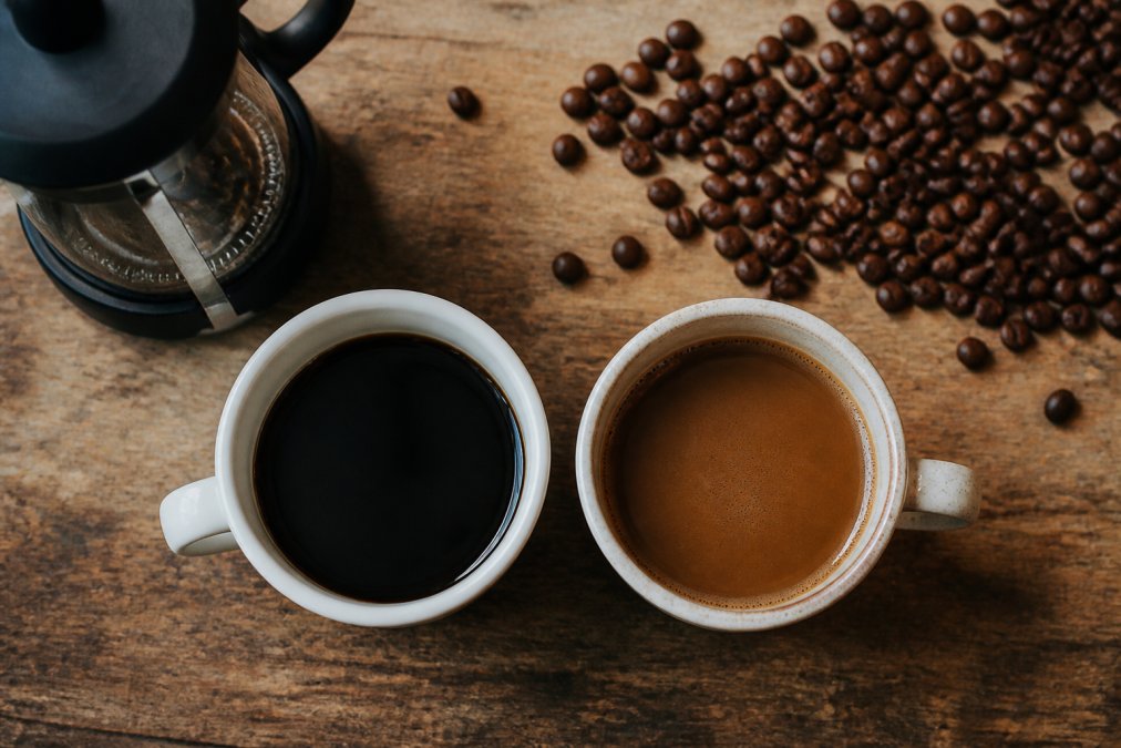 Overhead shot of two coffee cups side by side on a wooden kitchen counter, one made with dark supermarket coffee and one with lighter specialty coffee, with a cafetière and scattered coffee beans nearby