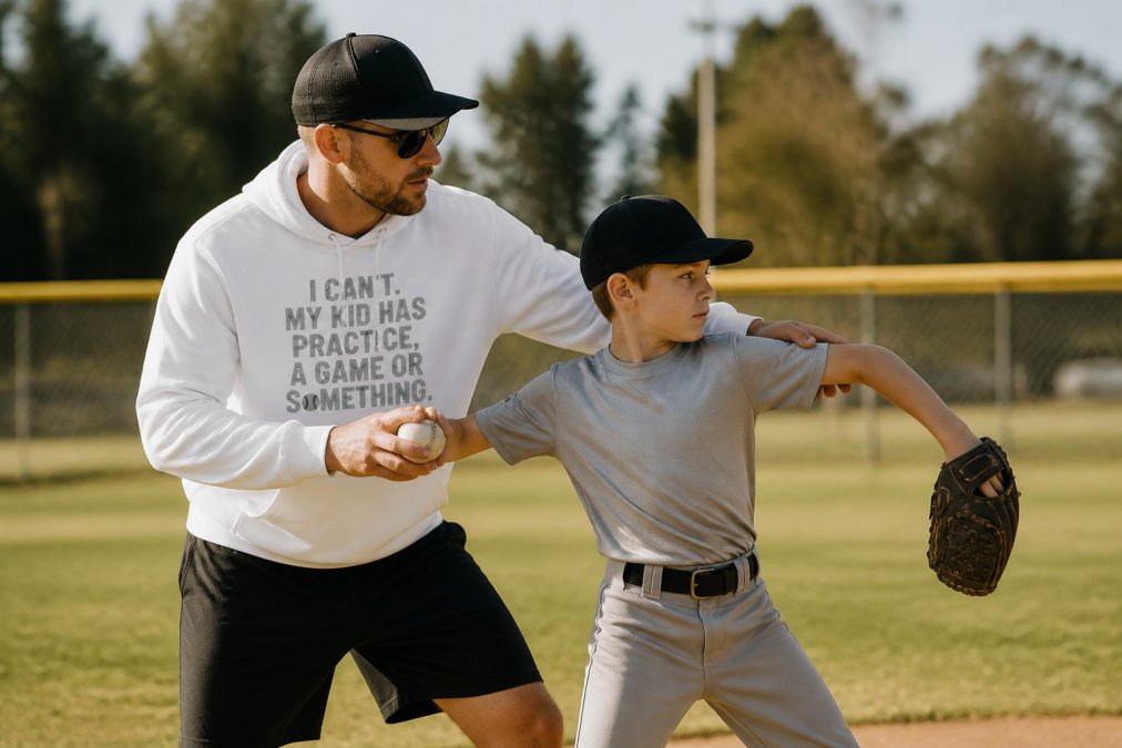 Youth coach demonstrating proper throwing technique with young baseball player outdoors on field during afternoon training session with natural sunlight