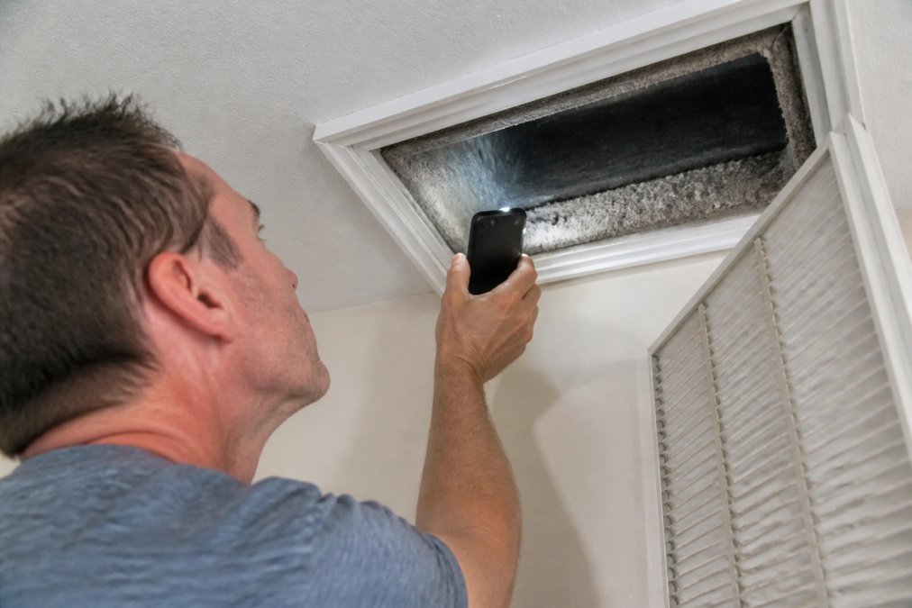 A close-up photo of a homeowner's hand holding a smartphone flashlight inside an open HVAC vent, revealing visible dust buildup and debris on the duct walls