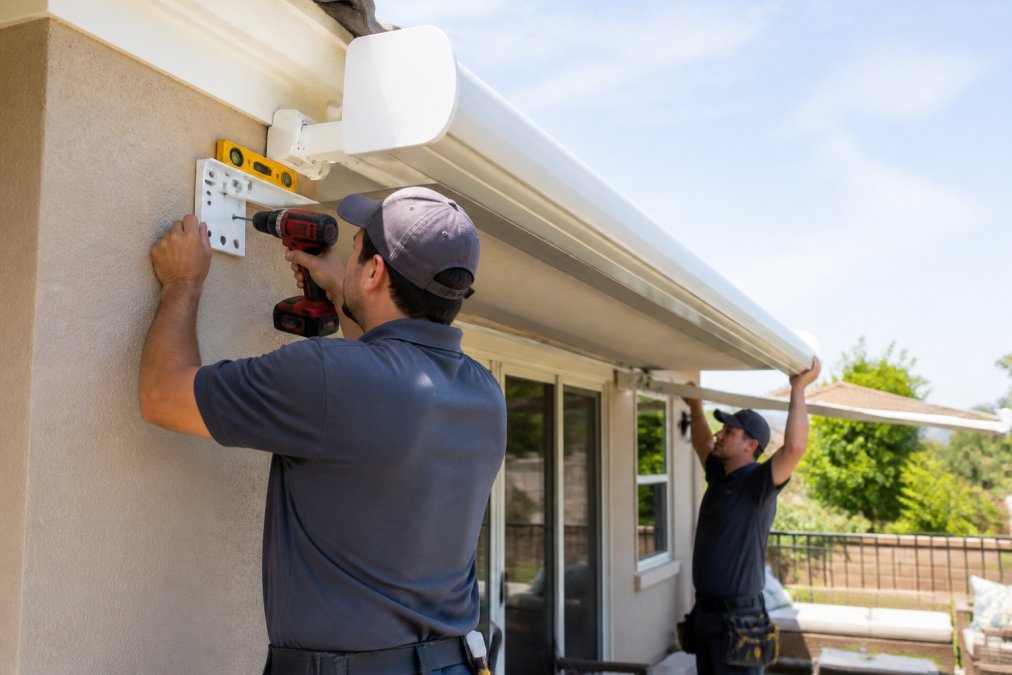 Professional installers using a drill and level for professional awning installation on a sunny patio wall.