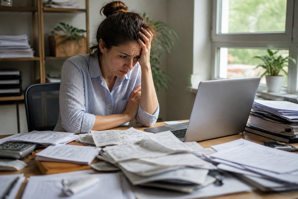 A small business owner sitting at a cluttered desk surrounded by piles of receipts and financial paperwork, looking at a laptop with a spreadsheet visible on the screen, stress visible in their posture, natural window light