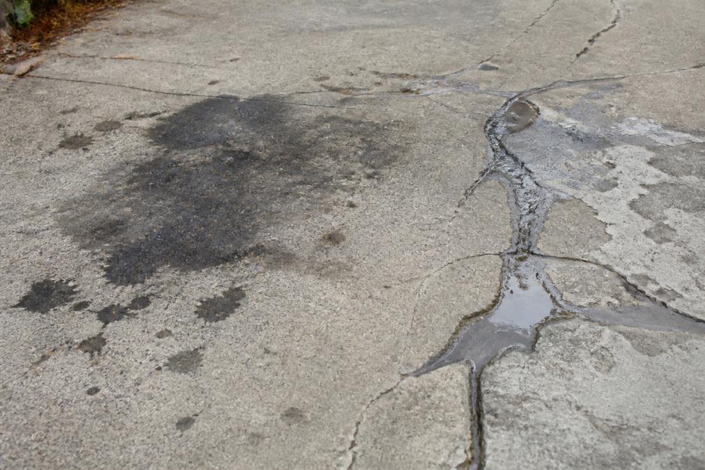 Close-up of concrete driveway surface showing dark stains from oil, visible cracks with water pooling, and faded areas where protective coating has worn away