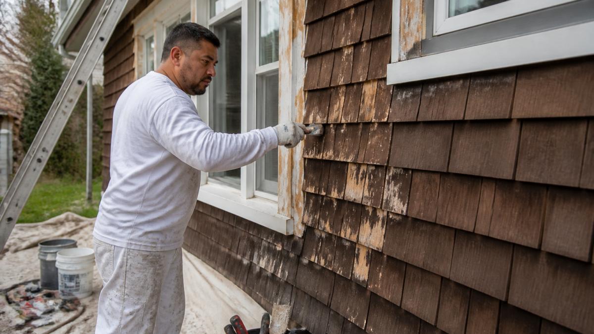 A professional painter in work clothes scraping and sanding a wood shingle exterior on a two-story home, with a ladder, drop cloths, and caulking tools visible, showing detailed surface preparation before painting