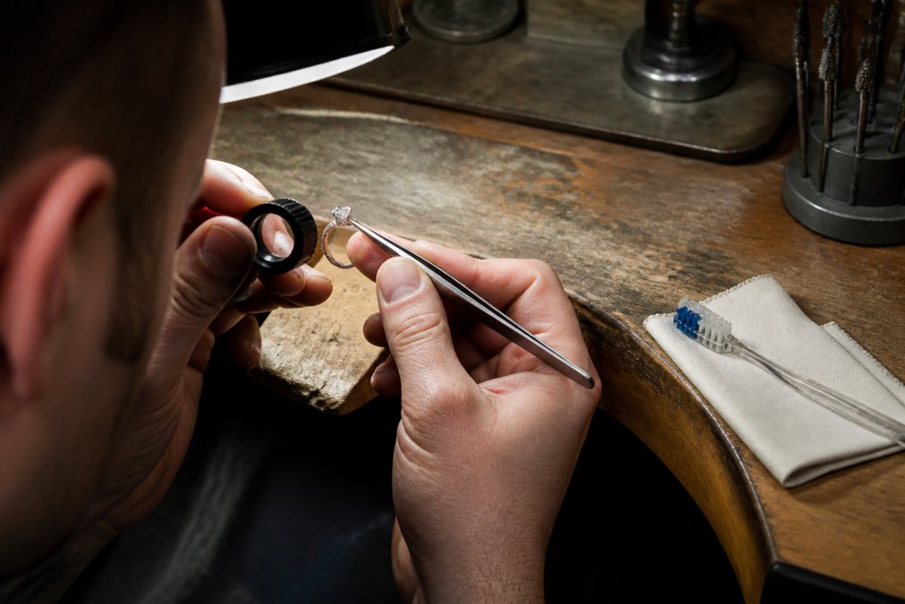 Jeweller using a loupe and tweezers to inspect a diamond ring setting on a workbench with cleaning tools nearby