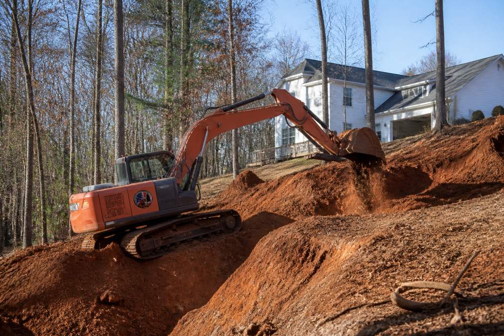 Professional grading equipment operating on residential property during active land clearing, showing heavy machinery working on steep terrain with visible soil displacement