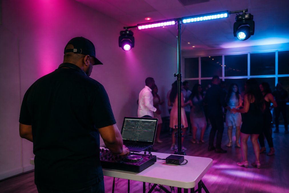 A mobile DJ performing at an evening event, standing behind a compact laptop-based lighting control setup on a folding table, colorful Govee smart light strips and two moving head fixtures mounted on a lightweight stand behind the DJ booth, casting pink and blue wash light across a small dance floor