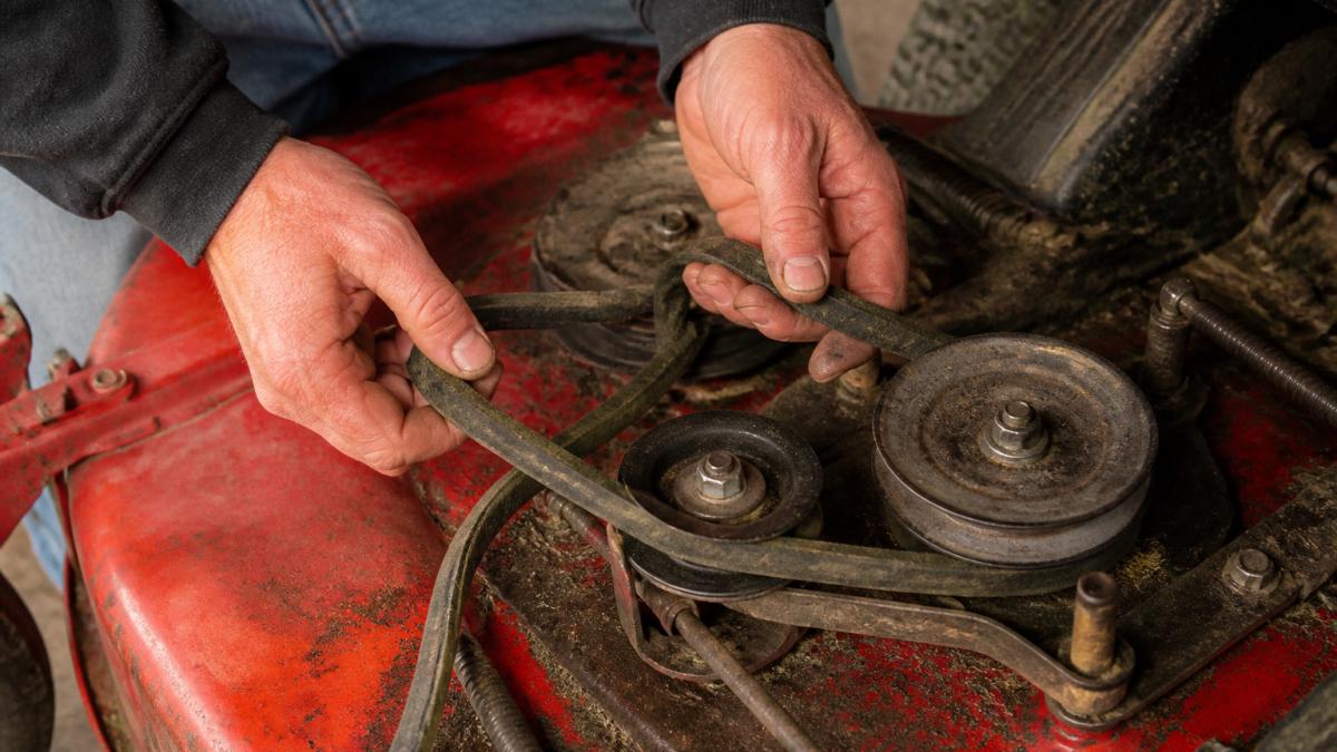 Close-up of mechanic's hands removing a worn mower deck belt from around pulleys and idler mechanisms, showing the belt routing path