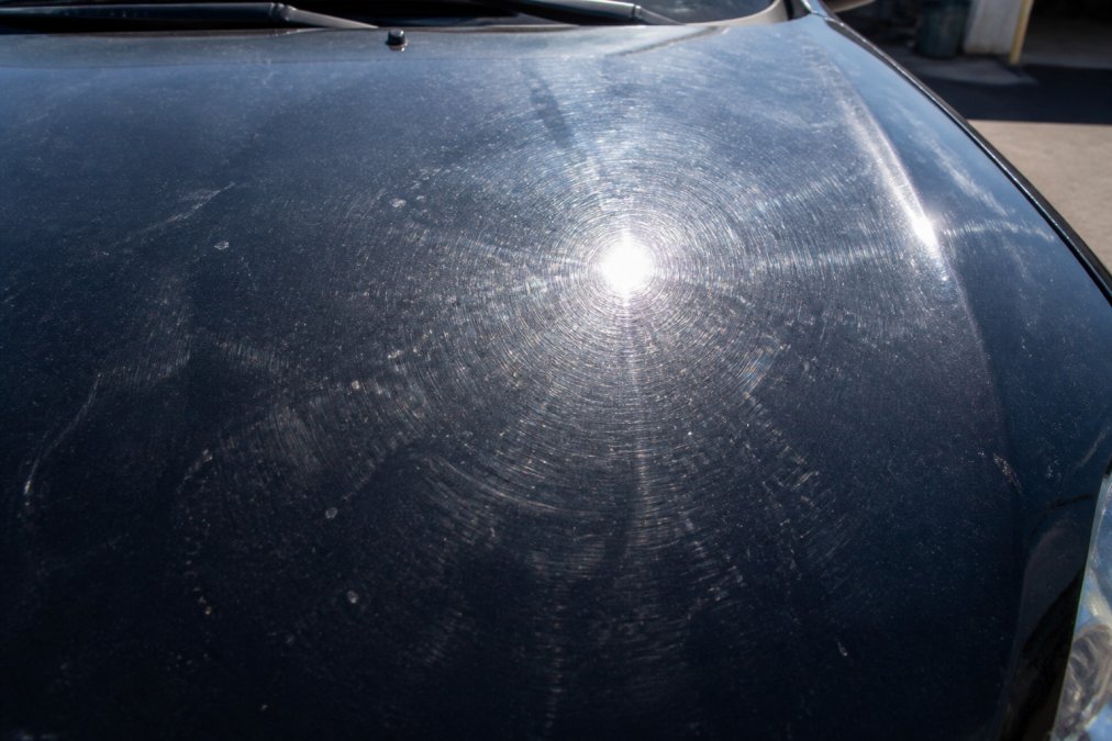 Close-up of vehicle hood showing visible swirl marks, oxidation spots, and surface contamination under natural daylight
