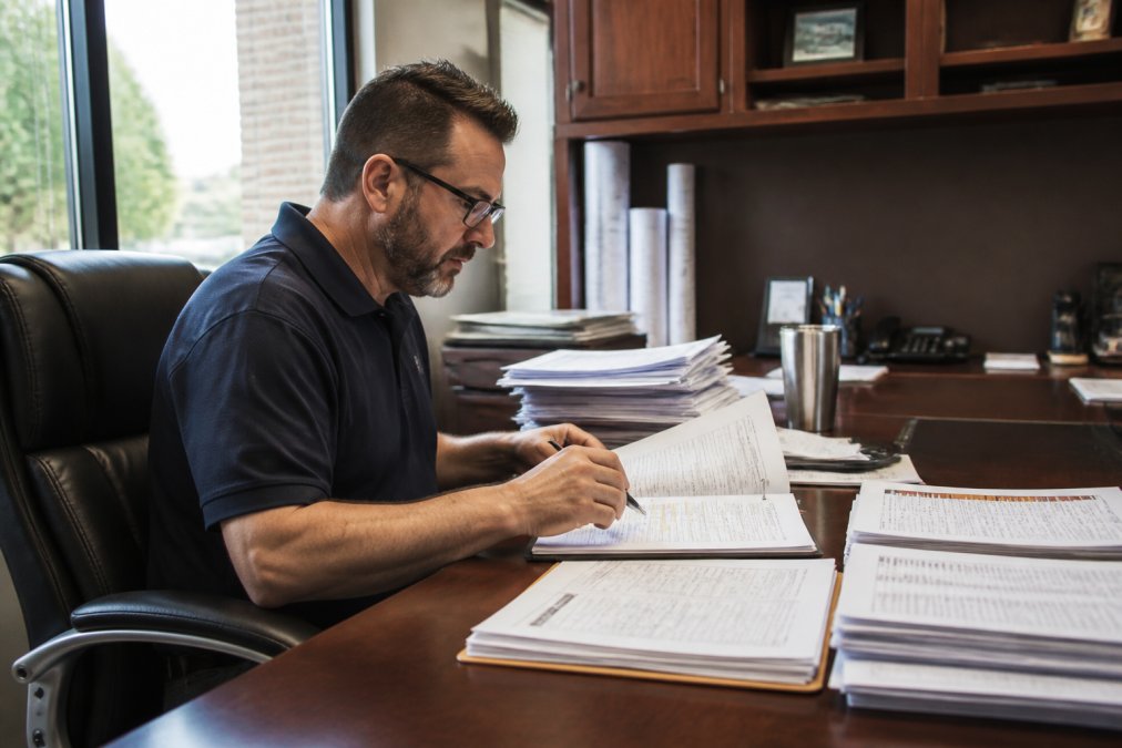 Project manager in office environment reviewing detailed budget spreadsheets and construction timelines on computer monitor with printed project documentation, permits, and cost reports visible on desk