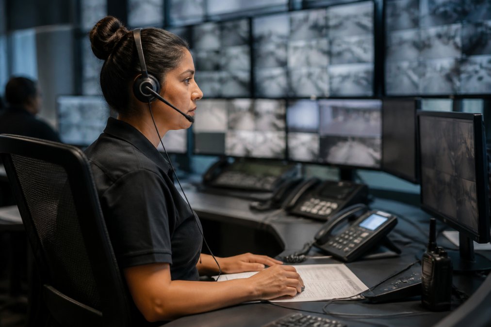 A security monitoring center operator wearing a headset, looking at multiple screens showing live camera feeds and alarm system status maps, with a calm, focused expression