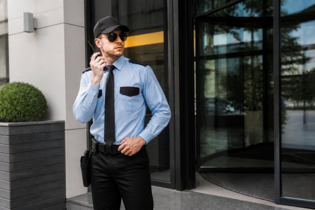A uniformed professional security guard standing at the entrance of a commercial building in daylight, radio in hand, scanning the car park with alert posture and high-visibility vest