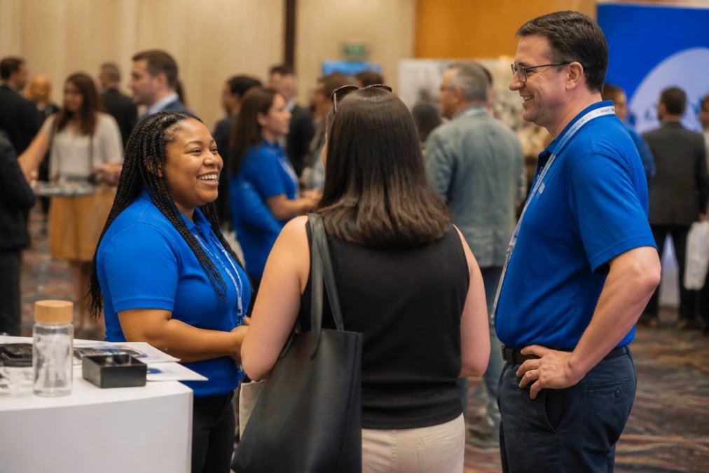 Professional photographer discreetly capturing candid moments as attendees interact with a sponsor booth, genuine smiles and engagement visible, natural event lighting
