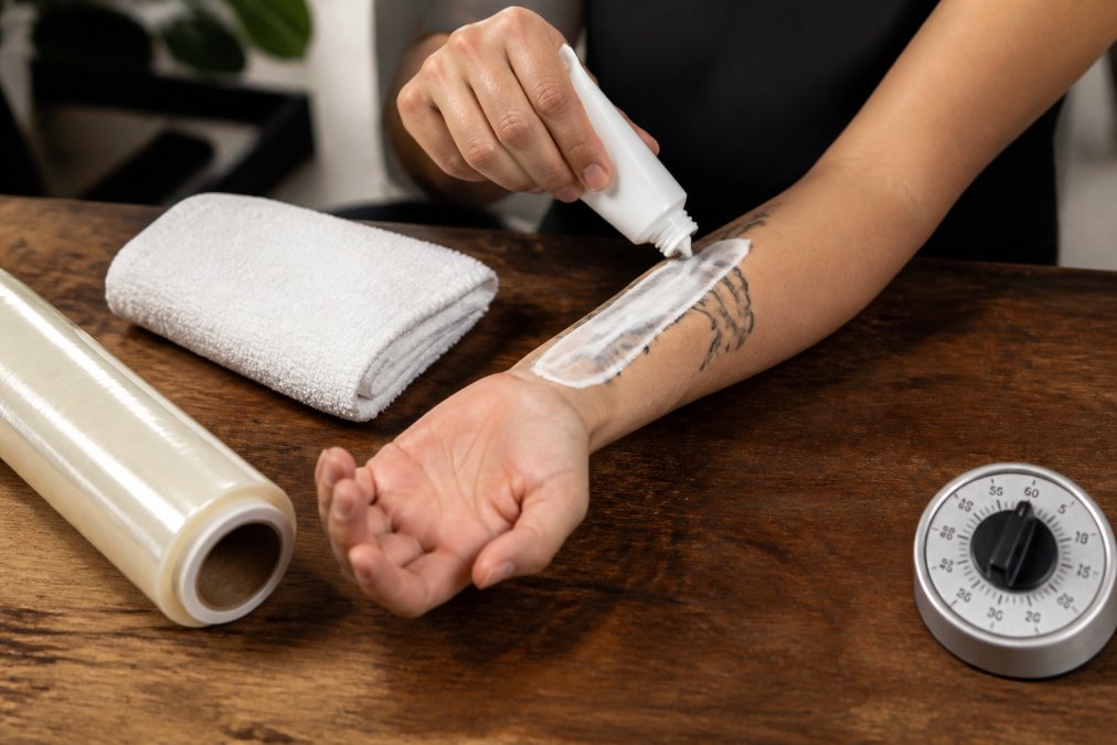 Close-up of hands applying white numbing cream from a tube onto a forearm, with a clean towel, plastic wrap, and a kitchen timer nearby on a wooden table