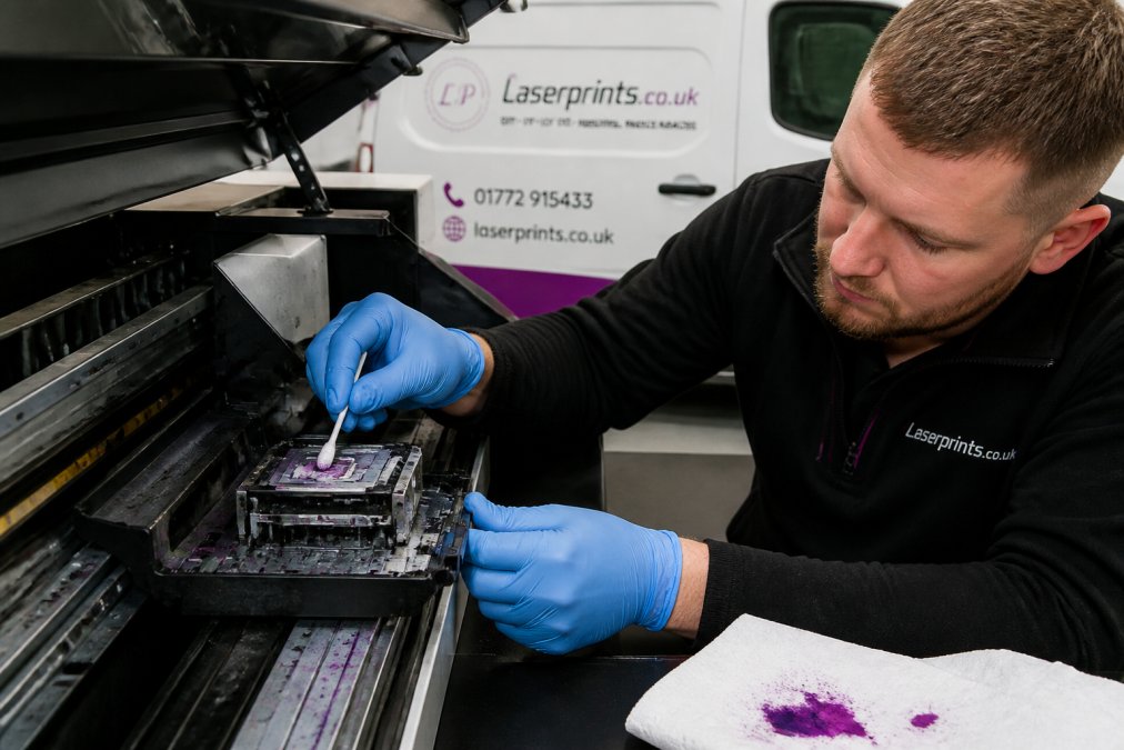 Close-up of a UV printer printhead being carefully cleaned by a technician wearing nitrile gloves, UV ink residue visible on a white cloth beside the machine in a well-lit workshop