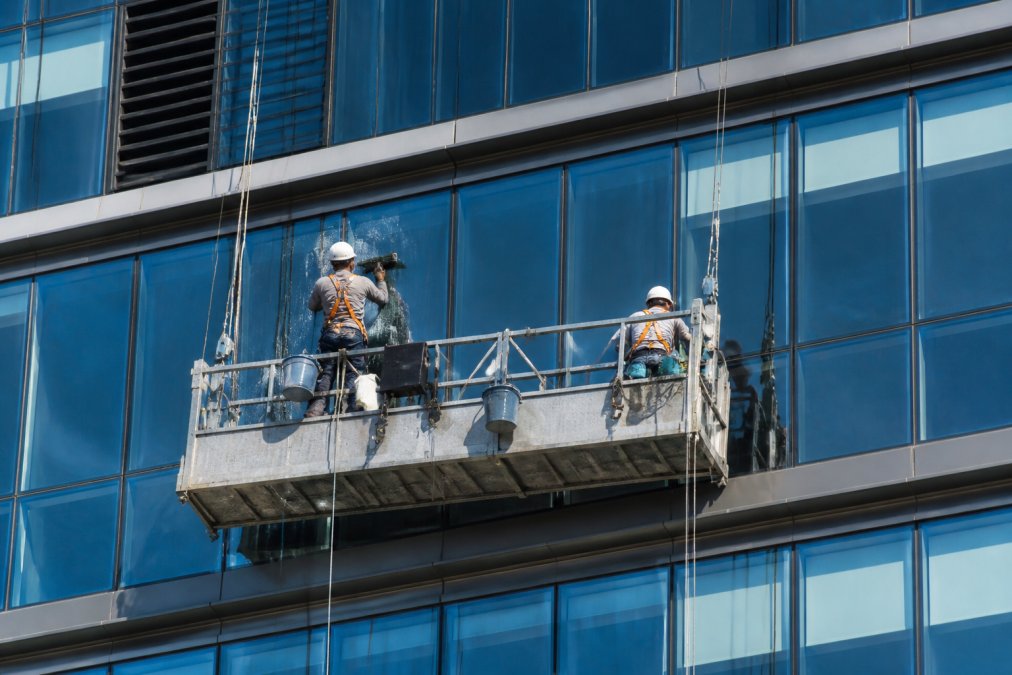 Two window cleaning technicians on a swing stage platform secured to anchor points on a high-rise building exterior, both wearing full personal protective equipment including helmets, harnesses, and gloves, working under bright daylight with the building's glass facade in the background