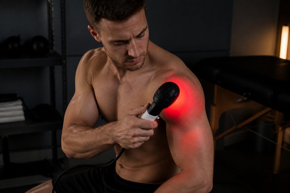 Athlete applying red light therapy device to shoulder muscle after workout, with red glow visible on skin in dimly lit recovery room