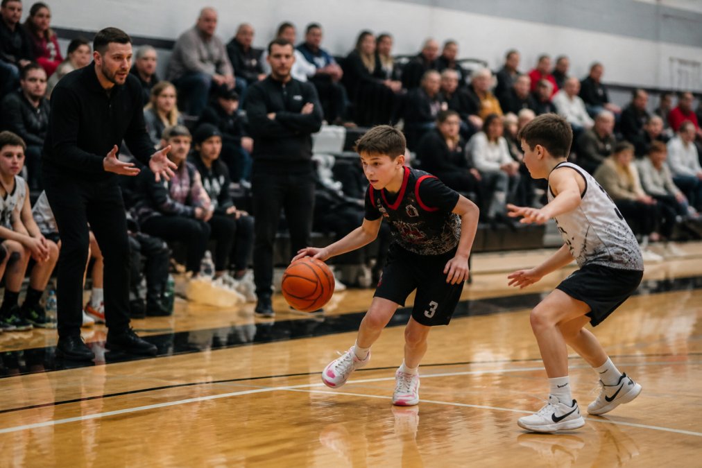 A youth basketball player dribbling past a defender during a competitive indoor game under gymnasium lights, with coaches gesturing from the sideline and parents watching from bleachers in the background