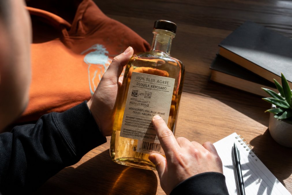 Close-up of a hand holding a reposado tequila bottle, pointing to the NOM number and ingredient list on the label with natural sunlight illuminating the text