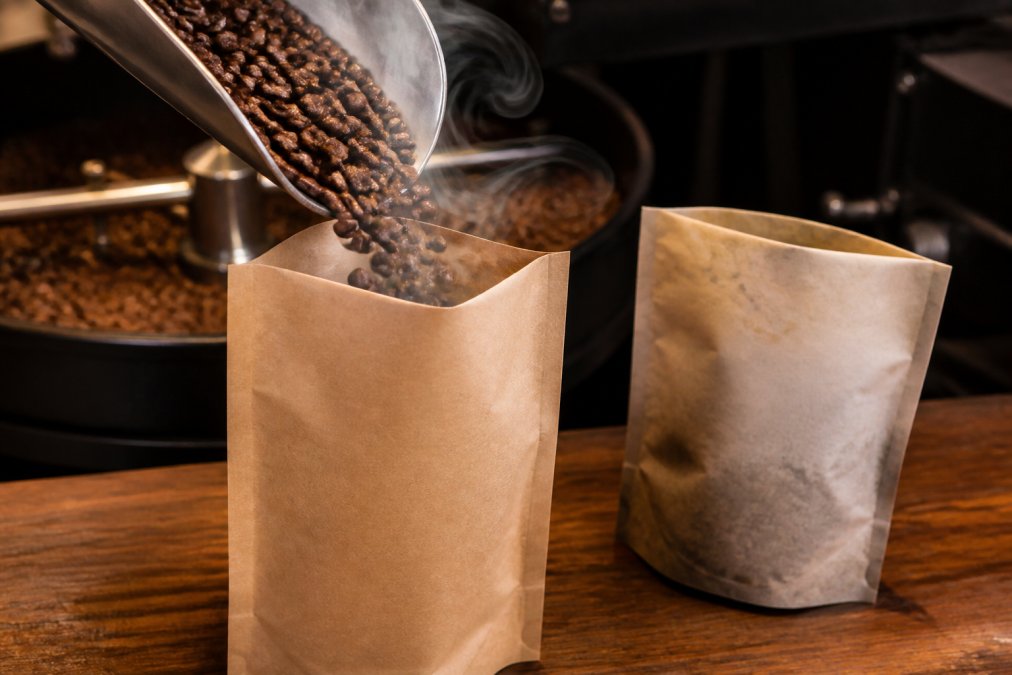 Close-up of freshly roasted coffee beans being poured into a kraft paper bag with visible steam rising, showing glossy dark beans with rich color, contrasted with a faded older coffee bag beside it on a wooden counter