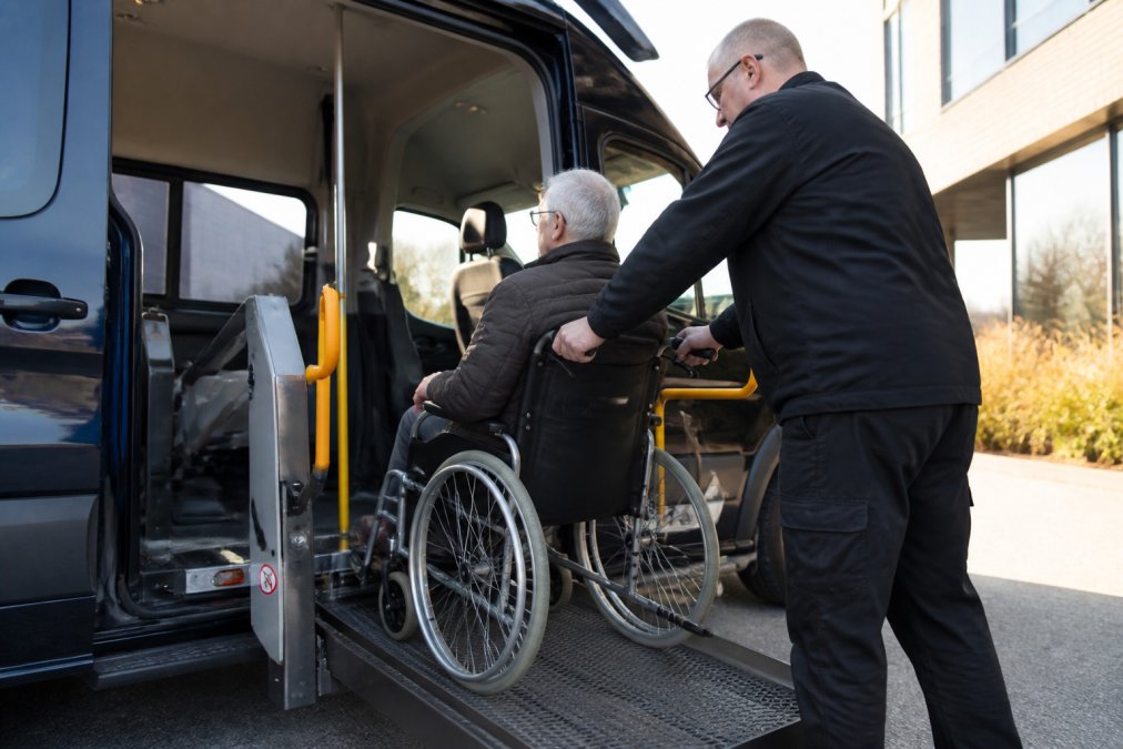 Professionele chauffeur helpt rolstoelgebruiker voorzichtig in een aangepast voertuig met zichtbare liftinstallatie, close-up van metalen beveiligingsbeugels en gordels, daglicht door de open zijdeur
