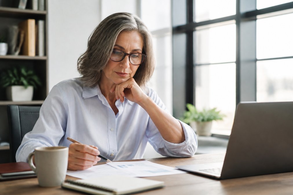 Professional woman in her 50s reviewing financial documents at a modern desk with laptop, coffee, and notepad, natural office lighting streaming through windows, focused and thoughtful expression