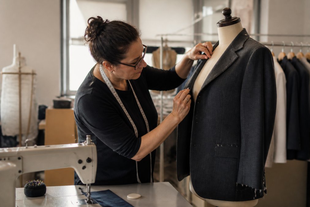 A seamstress at a sewing machine pinning a suit jacket on a dress form in a bright tailoring studio, measuring tape around her neck and chalk nearby