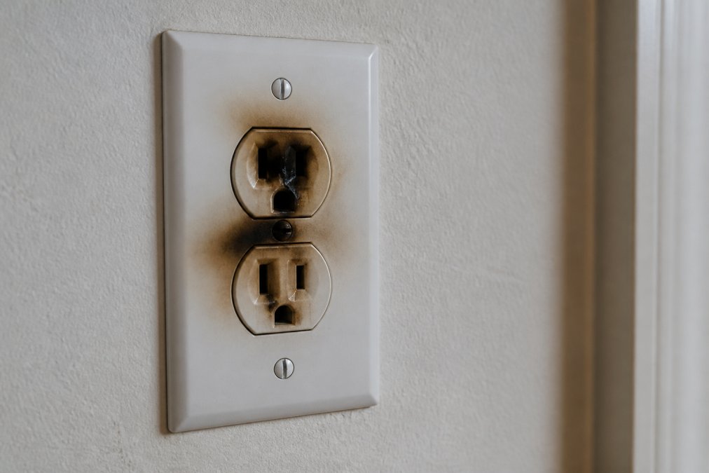 Close-up of a residential electrical outlet with visible burn marks and dark discoloration around the socket, showing signs of arcing damage and overheating on a white wall