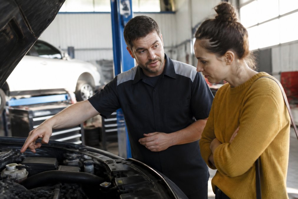 Professional automotive technician in shop uniform speaking with customer while pointing to vehicle's engine bay, demonstrating communication and transparency in a well-lit service facility