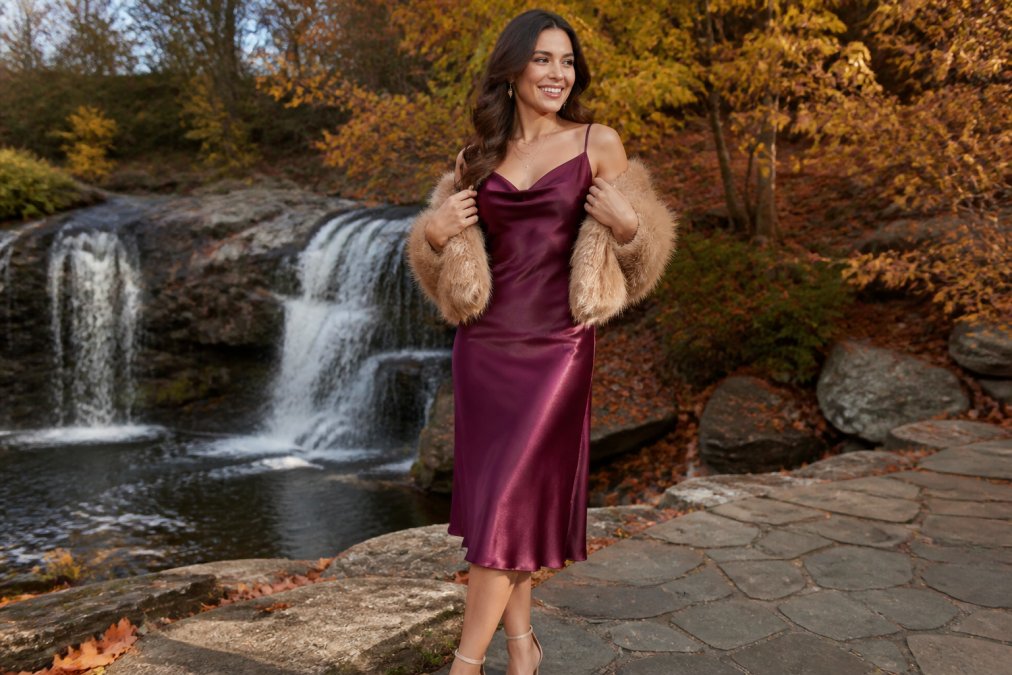 A woman in a rich plum satin midi dress standing outdoors among autumn foliage, warm afternoon sunlight catching the fabric's sheen, styled with a faux fur wrap draped over her shoulders