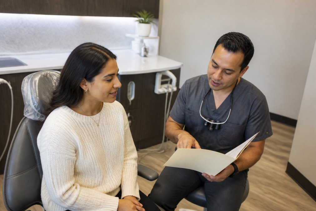 A patient in a dental chair speaking with a dentist who is reviewing treatment notes and discussing anxiety management and medical needs, with calm lighting and professional setting