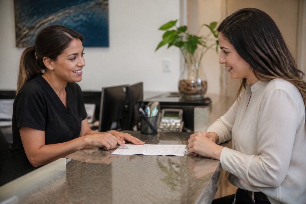 A patient at a dental office reception desk discussing payment plans with a friendly staff member, showing a collaborative conversation about billing and financing options with paperwork on the desk