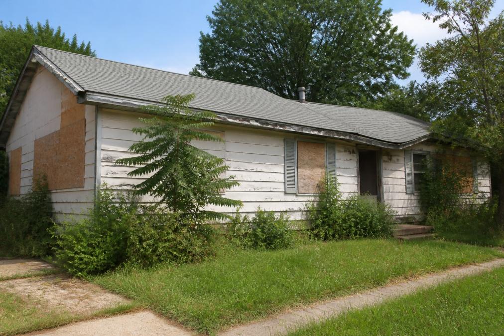 A single-story residential property with visible exterior damage including boarded windows, peeling paint, overgrown vegetation, and a sagging roof line photographed in natural daylight