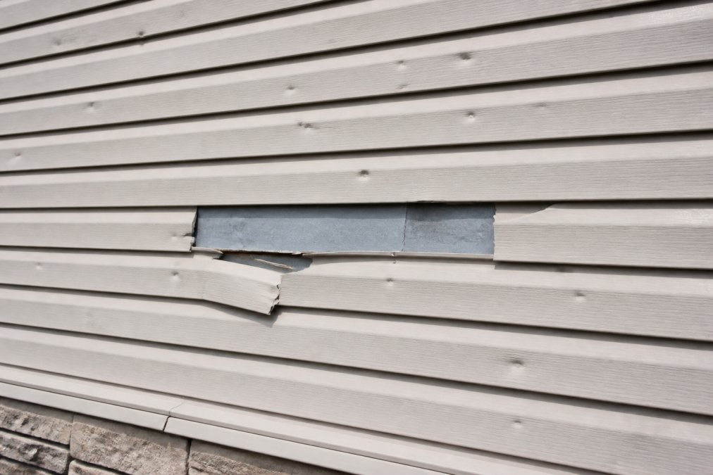 Close-up of residential home exterior showing siding with visible hail dents, cracks, and missing panels after severe storm damage