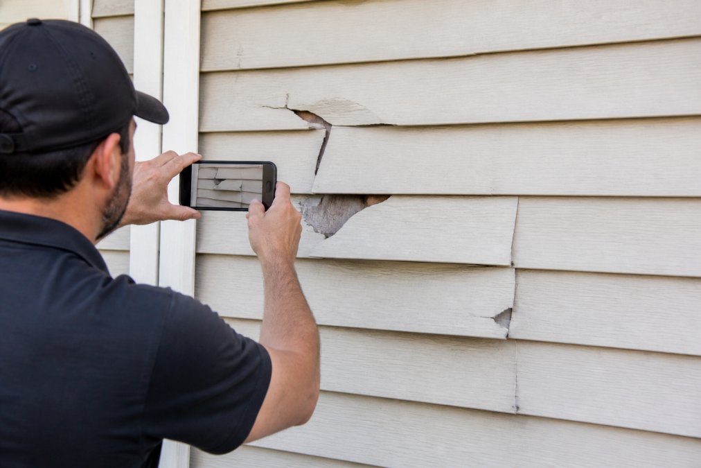 Homeowner photographing exterior siding damage with smartphone, capturing close-up details of cracks and missing panels in natural daylight for insurance documentation