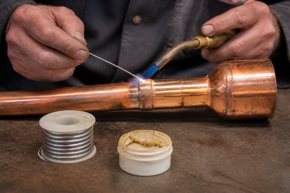 Close-up of a craftsman's hands using a soldering iron on a copper pipe joint, with a roll of silver solder and flux nearby on a workbench