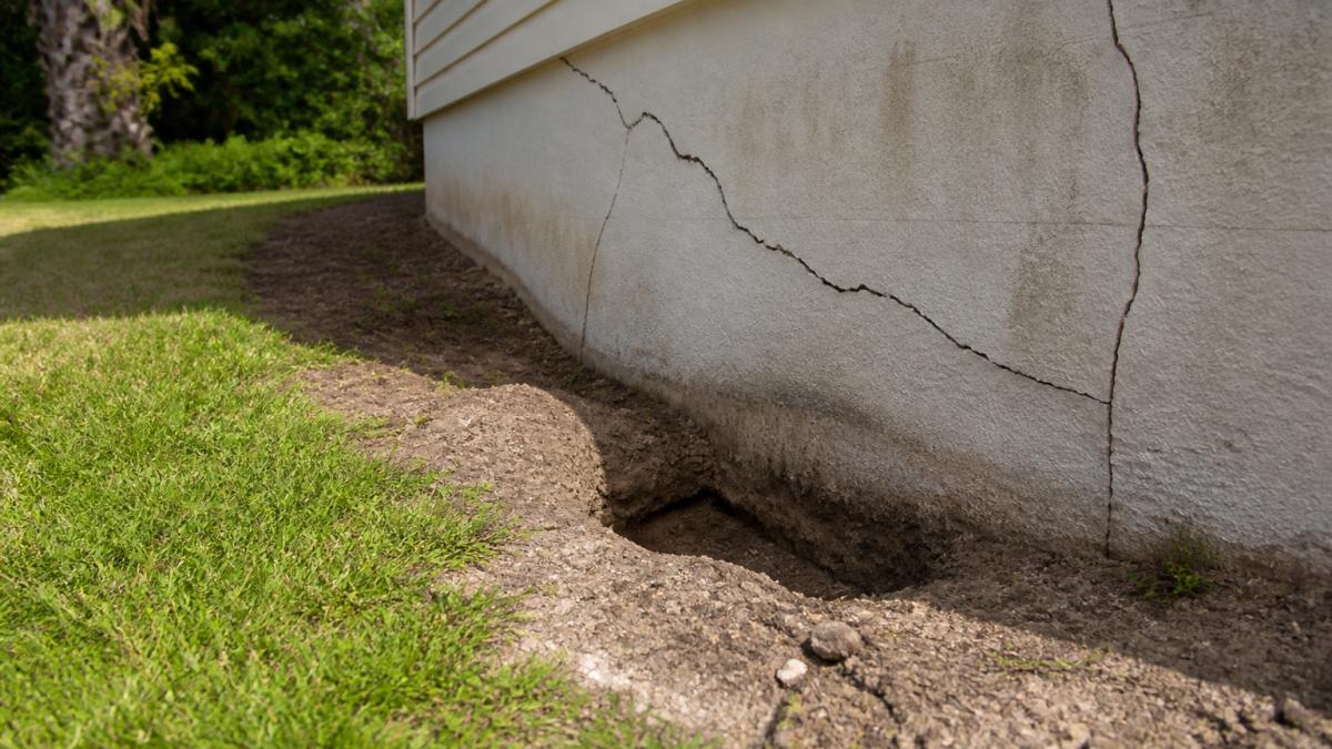 Close-up of diagonal foundation cracks and visible depression in soil around residential property foundation, showing structural damage and ground subsidence on sunny day