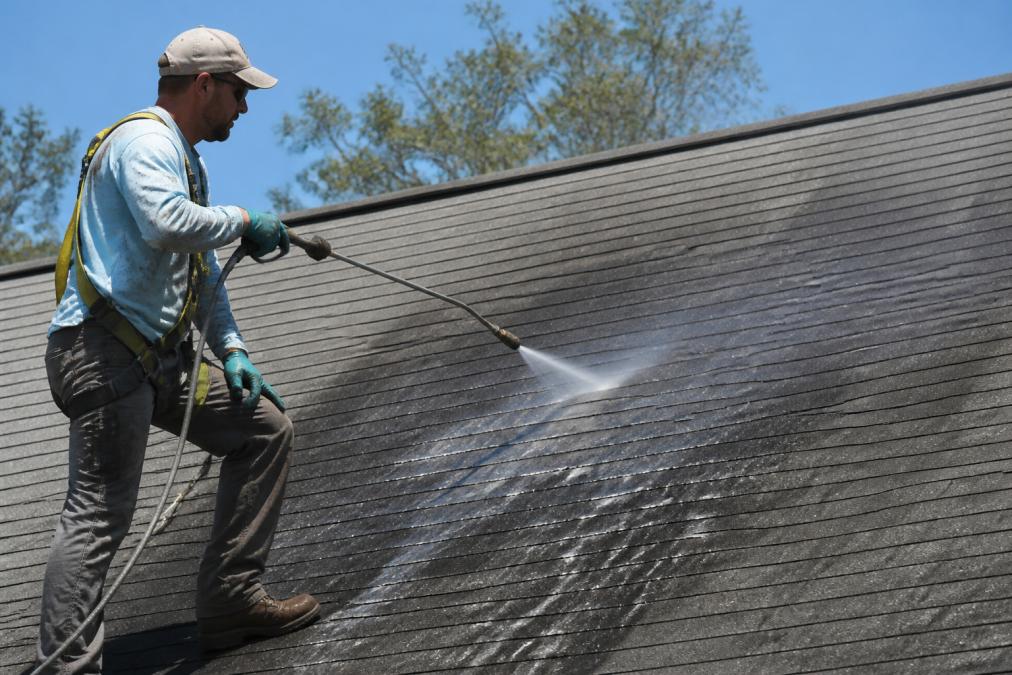 A technician in safety gear using a low-pressure soft wash wand on an asphalt shingle roof, with visible black algae streaks being rinsed away, set against a clear sky