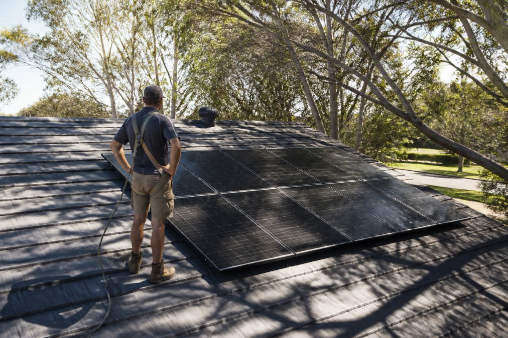 Homeowner standing on residential roof during afternoon with tree shadows visible across roof surface and solar panels partially shaded, natural daylight showing shade movement