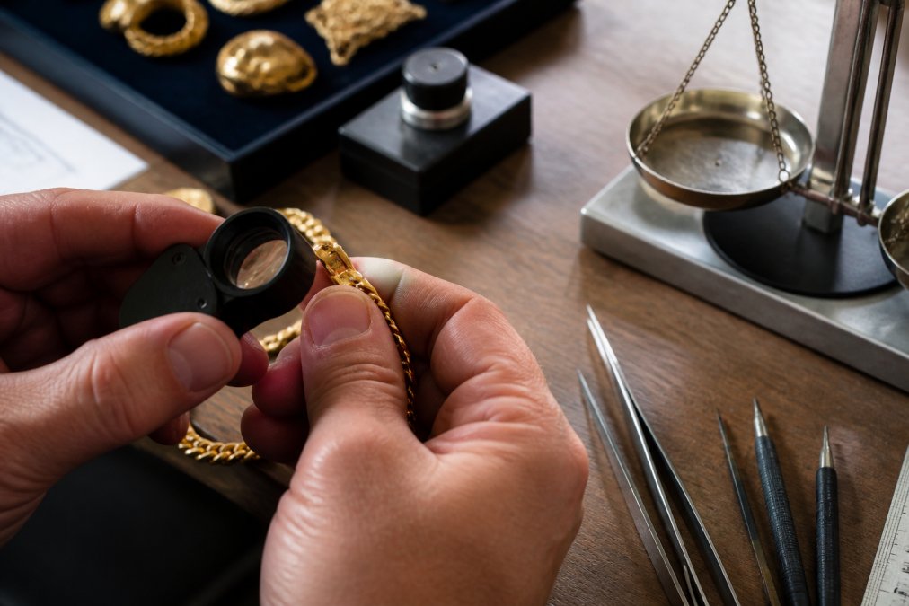 Close-up of jeweler's hands examining a gold necklace with a loupe, revealing detailed hallmark stamping on the metal surface under bright natural light