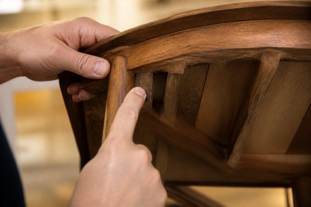 Close-up of hands inspecting the underside of a wooden chair, showing end grain and joinery details, warm indoor lighting